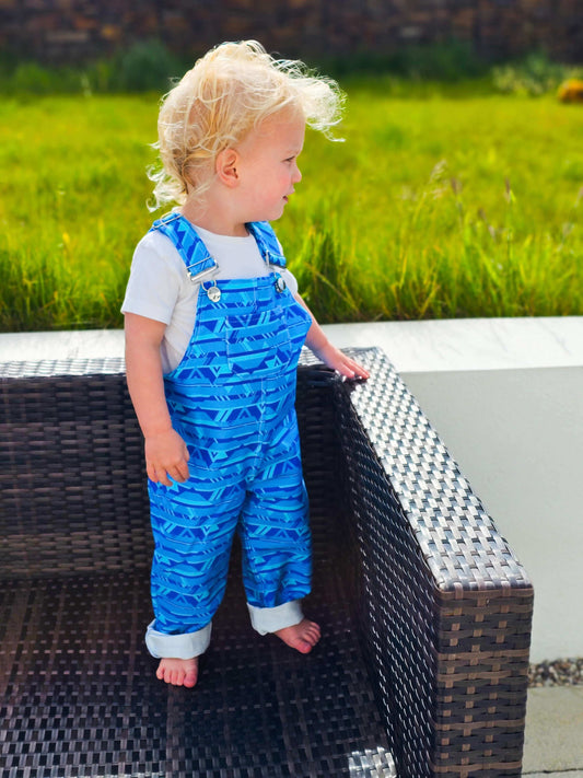 Toddler wearing blue handmade denim dungarees with adjustable straps and front pocket, standing outdoors on a wicker chair.